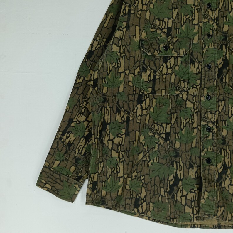 May include: A long-sleeved button-down shirt with a camouflage pattern. The shirt features a mix of brown, green, and black colors, with a leaf design. It has a chest pocket and black buttons.