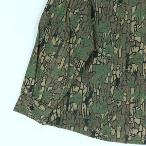 May include: Long-sleeved button-down shirt with a camouflage pattern. The shirt features a design of green leaves and brown tree bark. The cuff has a single black button.