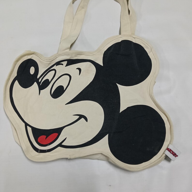 May include: A cream-colored tote bag shaped like the head of Mickey Mouse. The bag features a large black and white graphic of the character's face with a red tongue. Two fabric handles are attached to the top, and a small white tag with the text MICKEY is visible on the right side.