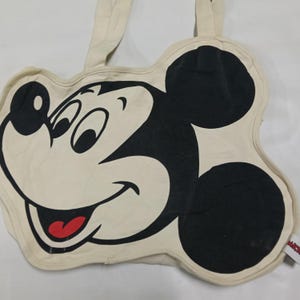 May include: A cream-colored canvas tote bag shaped like the head of Mickey Mouse. The bag features a large black graphic of the character's face with white eyes and a red tongue. Two fabric handles extend from the top of the shaped bag design.