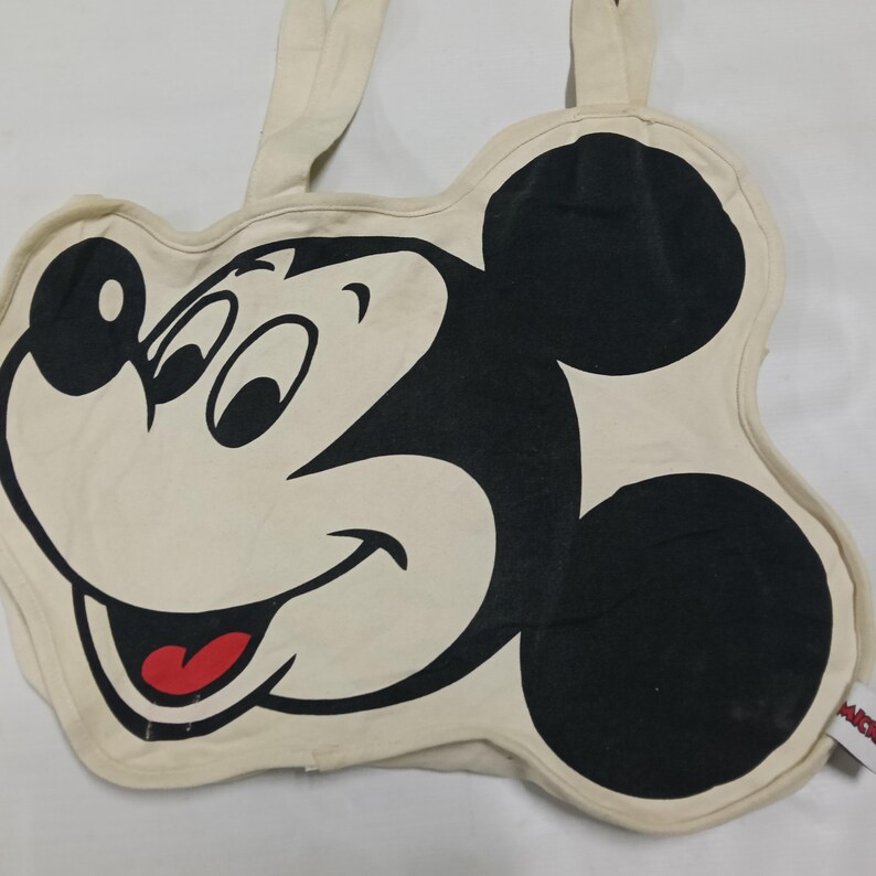 May include: A cream-colored canvas tote bag shaped like the head of Mickey Mouse. The bag features a large black graphic of the character's face with white eyes and a red tongue. Two fabric handles extend from the top of the shaped bag design.