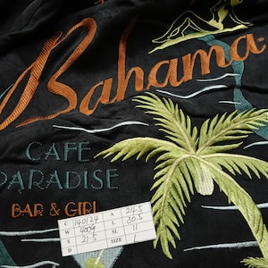 Peut inclure: Tissu noir avec broderie. Le motif comprend le mot "Bahama" en orange, un palmier et le texte "CAFE PARADISE BAR & GIRL". Une silhouette et des vagues sont &eacute;galement pr&eacute;sentes. Une &eacute;tiquette de taille est visible.