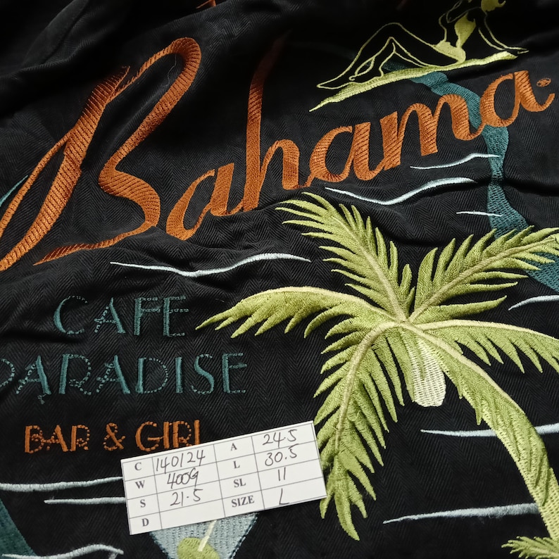 Peut inclure: Tissu noir avec broderie. Le motif comprend le mot "Bahama" en orange, un palmier et le texte "CAFE PARADISE BAR & GIRL". Une silhouette et des vagues sont &eacute;galement pr&eacute;sentes. Une &eacute;tiquette de taille est visible.