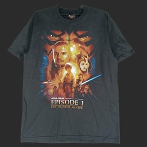 May include: Dark gray Star Wars Episode I t-shirt featuring a movie poster design. The graphic includes characters like Qui-Gon Jinn, Anakin Skywalker, and Queen Amidala, with the film's title prominently displayed.