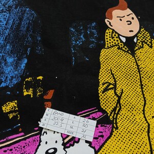 Puede incluir: Camiseta negra con un dise&ntilde;o gr&aacute;fico que presenta una ilustraci&oacute;n de dibujos animados de un personaje con abrigo amarillo y un perro blanco. El dise&ntilde;o tiene un estilo de c&oacute;mic con fondo negro y acentos en azul, amarillo y rosa. Una etiqueta blanca con medidas es visible.