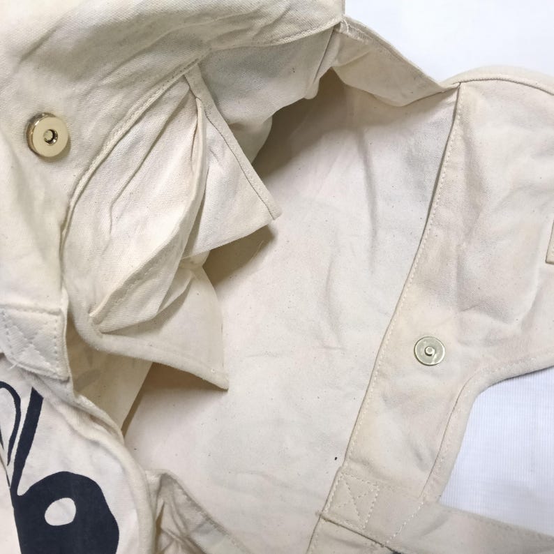May include: An off-white canvas tote bag interior featuring a large open main compartment, a side pocket, and a gold-toned magnetic snap closure. The fabric has a visible woven texture, and a portion of a dark graphic print is visible on the exterior.