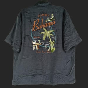 Peut inclure: Chemise grise fonc&eacute;e &agrave; manches courtes. Le dos pr&eacute;sente une broderie color&eacute;e avec les mots "Tommy Bahama" et "Cafe Paradise Bar & Grill". Le motif comprend un palmier, une femme et un cocktail.