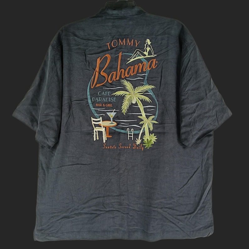Peut inclure: Chemise grise fonc&eacute;e &agrave; manches courtes. Le dos pr&eacute;sente une broderie color&eacute;e avec les mots "Tommy Bahama" et "Cafe Paradise Bar & Grill". Le motif comprend un palmier, une femme et un cocktail.