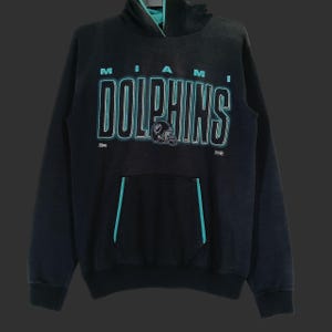 Könnte beinhalten: Schwarzer Kapuzenpullover mit dem Text "MIAMI DOLPHINS" in Weiß mit türkisfarbener Umrandung. Der Hoodie hat eine türkis gefütterte Kapuze und Tasche sowie eine Footballhelm-Grafik. Ein klassischer Sportartikel.