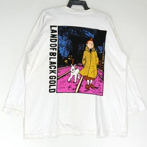 Puede incluir: Camiseta blanca de manga larga con un gr&aacute;fico de la serie de c&oacute;mics de Tint&iacute;n. El gr&aacute;fico muestra a Tint&iacute;n con un abrigo amarillo con Mil&uacute;, sobre un fondo rosa y morado con el texto "LAND OF BLACK GOLD".