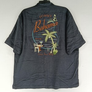 Peut inclure: Chemise boutonn&eacute;e gris fonc&eacute; avec un motif tropical au dos. Le motif comprend le texte "Tommy Bahama Cafe Paradise Bar & Grill" avec une femme, des palmiers et un cocktail. La chemise a des manches courtes.