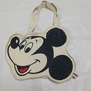 May include: A cream-colored canvas tote bag shaped like the head of Mickey Mouse. The bag features a large black and white graphic of the character's face with a red tongue. It includes two top handles and a small white tag with the text MICKEY.