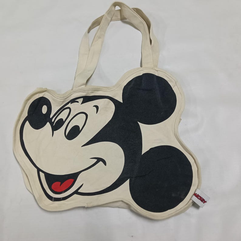 May include: A cream-colored canvas tote bag shaped like the head of Mickey Mouse. The bag features a large black and white graphic of the character's face with a red tongue. It includes two top handles and a small white tag with the text MICKEY.