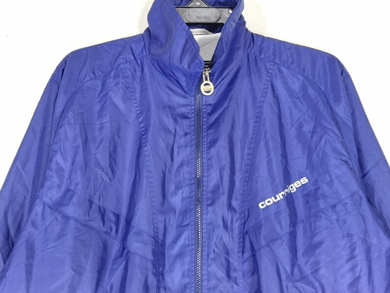 May include: A royal blue Courr&egrave;ges zip-up jacket. The jacket has a high collar and a silver zipper. The Courr&egrave;ges logo is printed in white on the front. The jacket is made of a lightweight, possibly nylon, material.