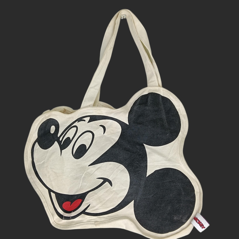 May include: A cream-colored canvas tote bag shaped like the head of Mickey Mouse. The bag features a large black graphic of the character's face with a red tongue and two top handles. A small white tag with the word Mickey is attached to the side.
