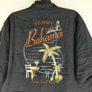 Peut inclure: Chemise boutonn&eacute;e gris fonc&eacute; avec un motif brod&eacute; au dos. Le motif pr&eacute;sente les mots "Tommy Bahama" en &eacute;criture orange, une femme, des palmiers et le texte "Cafe Paradise Bar & Girl".