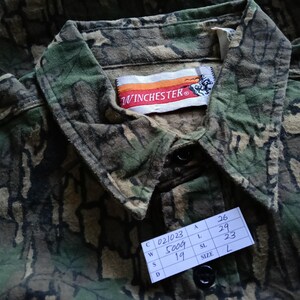 May include: Close-up of a camouflage button-down shirt with the "WINCHESTER" logo on the label. The shirt features a green, brown, and black pattern. A tag with the size "L" is visible.