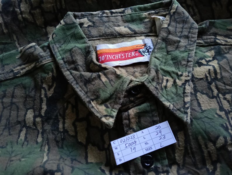 May include: Close-up of a camouflage button-down shirt with the "WINCHESTER" logo on the label. The shirt features a green, brown, and black pattern. A tag with the size "L" is visible.