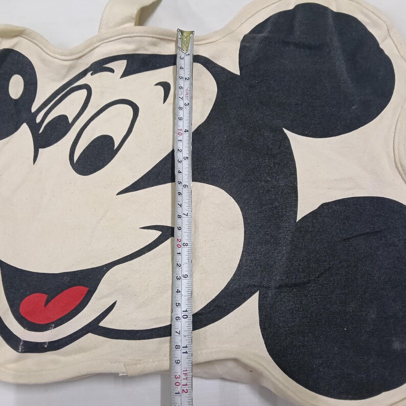 May include: A cream-colored canvas tote bag shaped like the head of Mickey Mouse, featuring a large black graphic of the character's face with a red tongue. A measuring tape is placed vertically across the center, showing a length of 12 inches.