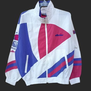 May include: White, blue, and pink vintage track jacket with a full zipper. The jacket features a geometric design with the brand name "ellesse" in blue. The jacket has a high collar and elastic cuffs.