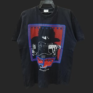 May include: Black t-shirt featuring a graphic of a musician playing a bass guitar. The design is framed in red and blue, with the text "THE BAD DOKER" at the top and "FIZZY" at the bottom. The shirt has a vintage look.