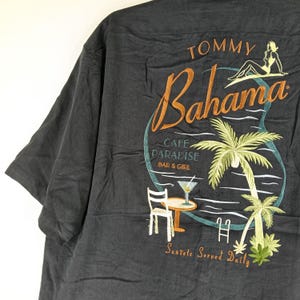 Peut inclure: Chemise Tommy Bahama gris fonc&eacute; avec un motif brod&eacute; au dos. Le motif pr&eacute;sente le mot "Bahama" en &eacute;criture orange, un palmier et les mots "Cafe Paradise Bar & Girl".