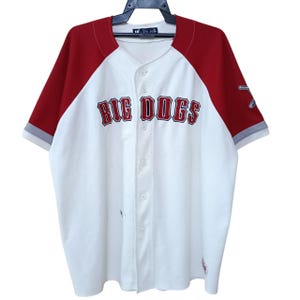 May include: White baseball jersey with red sleeves and collar. The text "BIG DOGS" is across the chest in a red, outlined font. The sleeves have grey trim. The jersey is button-down and has a tag at the neckline.