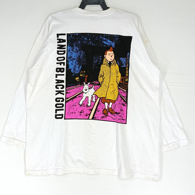 Puede incluir: Camiseta blanca de manga larga con un gr&aacute;fico de la serie de c&oacute;mics de Tint&iacute;n. El gr&aacute;fico muestra a Tint&iacute;n y Mil&uacute; caminando sobre una v&iacute;a de tren. El texto "LAND OF BLACK GOLD" est&aacute; impreso verticalmente en el lado izquierdo.