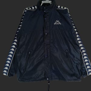 Peut inclure: Veste Kappa bleu marine avec des détails blancs. La veste a un col montant, une fermeture boutonnée et deux poches zippées. Logos Kappa blancs sur les manches et la poitrine. Cordon de serrage en bas.
