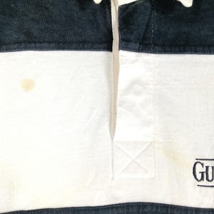 May include: Close-up of a white and navy blue rugby shirt. The shirt has a white body with navy blue sections at the top and bottom. The shirt has a small logo on the bottom right. There is a stain on the white section.