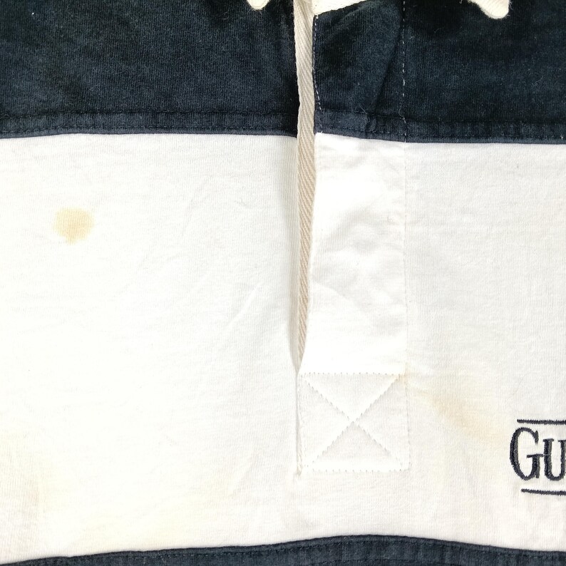 May include: Close-up of a white and navy blue rugby shirt. The shirt has a white body with navy blue sections at the top and bottom. The shirt has a small logo on the bottom right. There is a stain on the white section.
