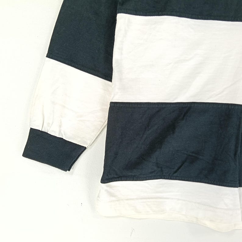 May include: Close-up of a long-sleeved shirt with a color block design. The shirt features alternating horizontal stripes of white and dark teal. The sleeve cuff and a section of the shoulder are dark teal.