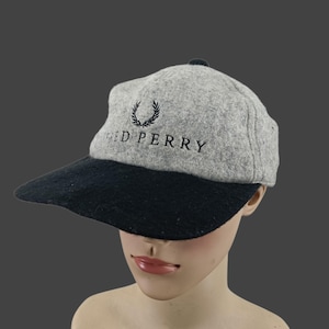May include: A gray and black baseball cap with the text "FRED PERRY" embroidered in black. The cap features a black brim and a small black button on top. The Fred Perry logo is above the text.