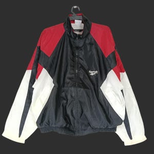 Op de afbeelding: Een vintage Reebok trainingsjack in zwart, rood en wit. De jas heeft een volledige ritssluiting, een opstaande kraag en het Reebok-logo op de borst. Het colorblock-ontwerp omvat rode schouders, een zwart midden en witte mouwen.