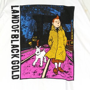 Puede incluir: Camiseta blanca con un dise&ntilde;o gr&aacute;fico de la historieta "Las aventuras de Tint&iacute;n". El dise&ntilde;o muestra a Tint&iacute;n con un abrigo amarillo, su perro Mil&uacute;, y el texto "LAND OF BLACK GOLD" en vertical. El fondo es rosa y azul.