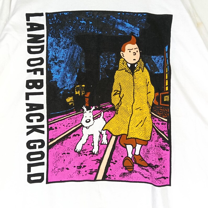Puede incluir: Camiseta blanca con un dise&ntilde;o gr&aacute;fico de la historieta "Las aventuras de Tint&iacute;n". El dise&ntilde;o muestra a Tint&iacute;n con un abrigo amarillo, su perro Mil&uacute;, y el texto "LAND OF BLACK GOLD" en vertical. El fondo es rosa y azul.