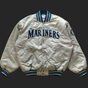 May include: A beige satin Mariners baseball jacket with teal and white striped trim on the collar, cuffs, and hem. The team name is embroidered in teal across the chest. A team logo patch is on the upper sleeve.