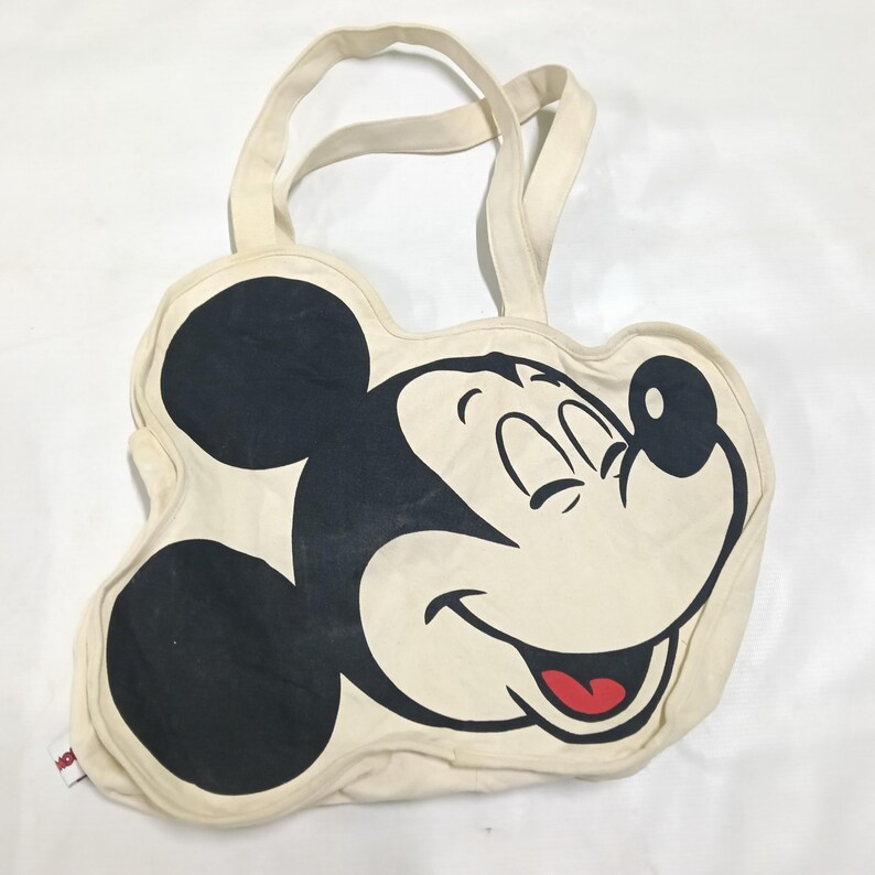 May include: A cream-colored canvas tote bag shaped like the head of Mickey Mouse. The bag features a large black graphic of Mickey Mouse's face with a smiling expression, a black nose, and a red tongue. It includes two matching cream-colored handles.