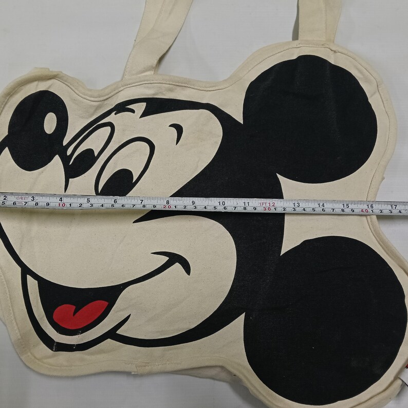 May include: A cream-colored canvas tote bag shaped like Mickey Mouse's head, featuring a large black and white graphic of his face with a red tongue. A measuring tape across the center shows a width of approximately 17 inches.