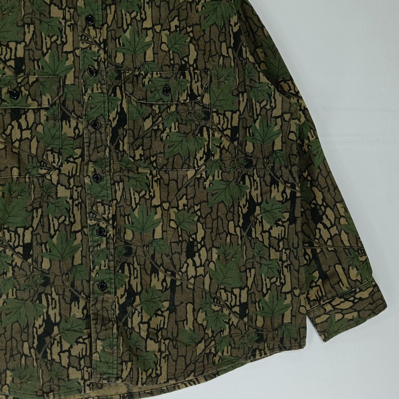 May include: A long-sleeved button-down shirt with a camouflage pattern. The shirt features a repeating design of green maple leaves and brown bark-like textures. It has two chest pockets and a button-down front.
