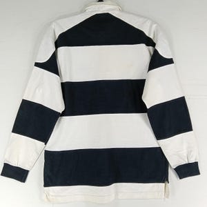 May include: A long-sleeved rugby shirt with alternating horizontal stripes of white and navy blue. The shirt has a white collar and cuffs, and a small zipper at the bottom hem. The shirt is made of a thick, durable fabric.
