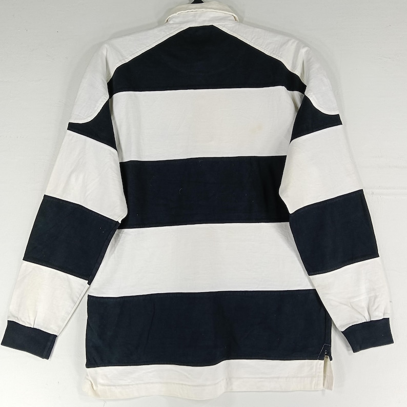 May include: A long-sleeved rugby shirt with alternating horizontal stripes of white and navy blue. The shirt has a white collar and cuffs, and a small zipper at the bottom hem. The shirt is made of a thick, durable fabric.