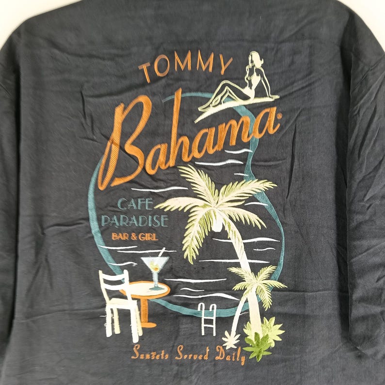 Peut inclure: T-shirt Tommy Bahama gris fonc&eacute; avec un motif brod&eacute;. Le motif comprend le mot "Bahama" en &eacute;criture orange, un palmier, une femme et les mots "Cafe Paradise Bar & Girl". Le texte "Sunsets Served Daily" est &eacute;galement inclus.