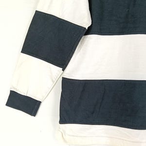 May include: A long-sleeved shirt with a color block design. The shirt features alternating horizontal stripes of white and navy blue. The sleeve has a white and navy blue color block pattern.