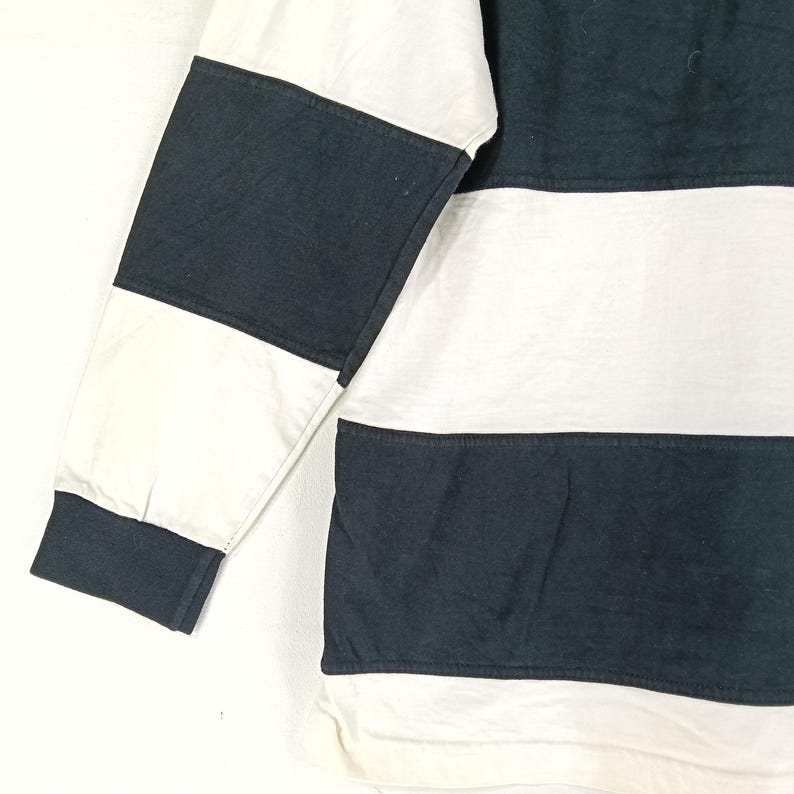 May include: A long-sleeved shirt with a color block design. The shirt features alternating horizontal stripes of white and navy blue. The sleeve has a white and navy blue color block pattern.