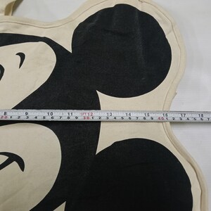 May include: A cream-colored canvas tote bag featuring a large black graphic of a cartoon mouse head. A measuring tape is placed across the center, showing a width of approximately 11 inches from the edge of the graphic to the right side.