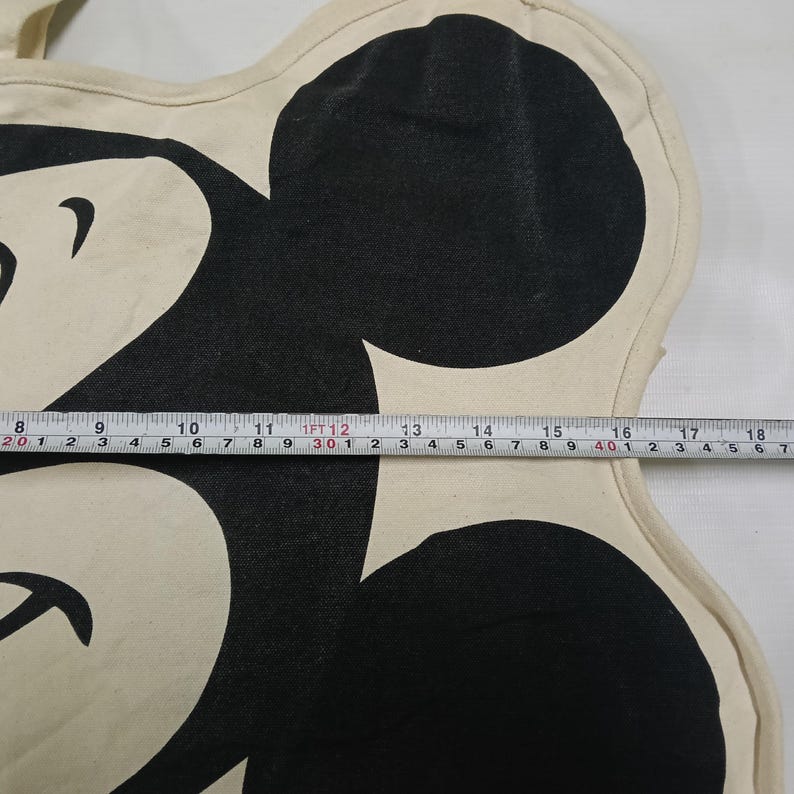 May include: A cream-colored canvas tote bag featuring a large black graphic of a cartoon mouse head. A measuring tape is placed across the center, showing a width of approximately 11 inches from the edge of the graphic to the right side.