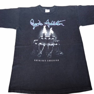 May include: Black t-shirt featuring a white graphic of two figures seated on a throne with flames above their heads. The text "Jane's Addiction" and "Nothing's Shocking" is printed on the shirt.