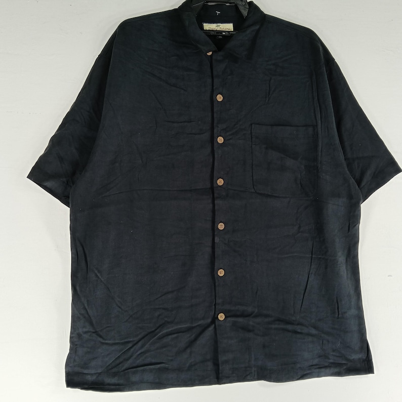 Peut inclure: Chemise noire &agrave; manches courtes avec boutons et une poche poitrine. La chemise a un col classique et des boutons de couleur bois. Le tissu semble &ecirc;tre l&eacute;ger, id&eacute;al pour les climats chauds.
