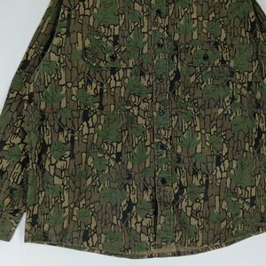 May include: A long-sleeved button-down shirt with a camouflage pattern featuring green maple leaves and a brown bark-like background. The shirt has two chest pockets and black buttons. The fabric appears to be a medium-weight material.